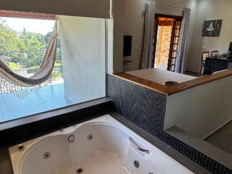 Uma banheira de hidromassagem branca fica ao lado de uma grande janela com vista para uma paisagem verdejante e exuberante, com uma rede em um pátio.