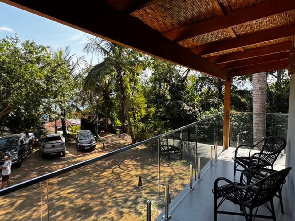 Vista da varanda com cadeiras escuras, guarda-corpo de vidro e cobertura de madeira sombreada, com vista para um estacionamento e árvores.