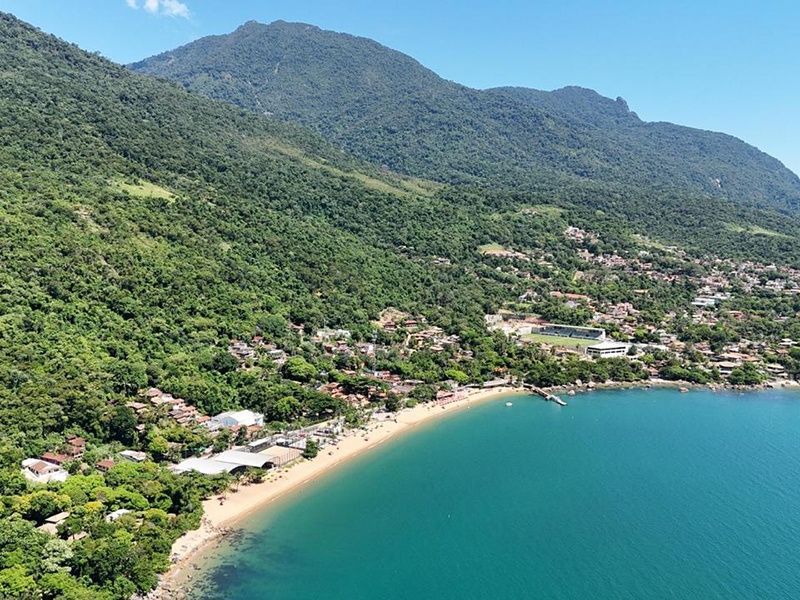 Vista aérea de uma praia de areia aninhada contra uma montanha verdejante, com água cristalina turquesa em primeiro plano.