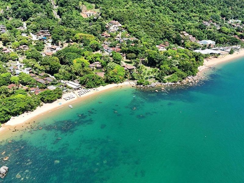 Uma vista aérea de uma praia de areia que encontra árvores verdejantes e casas na encosta, margeando as águas cristalinas e turquesas do oceano.