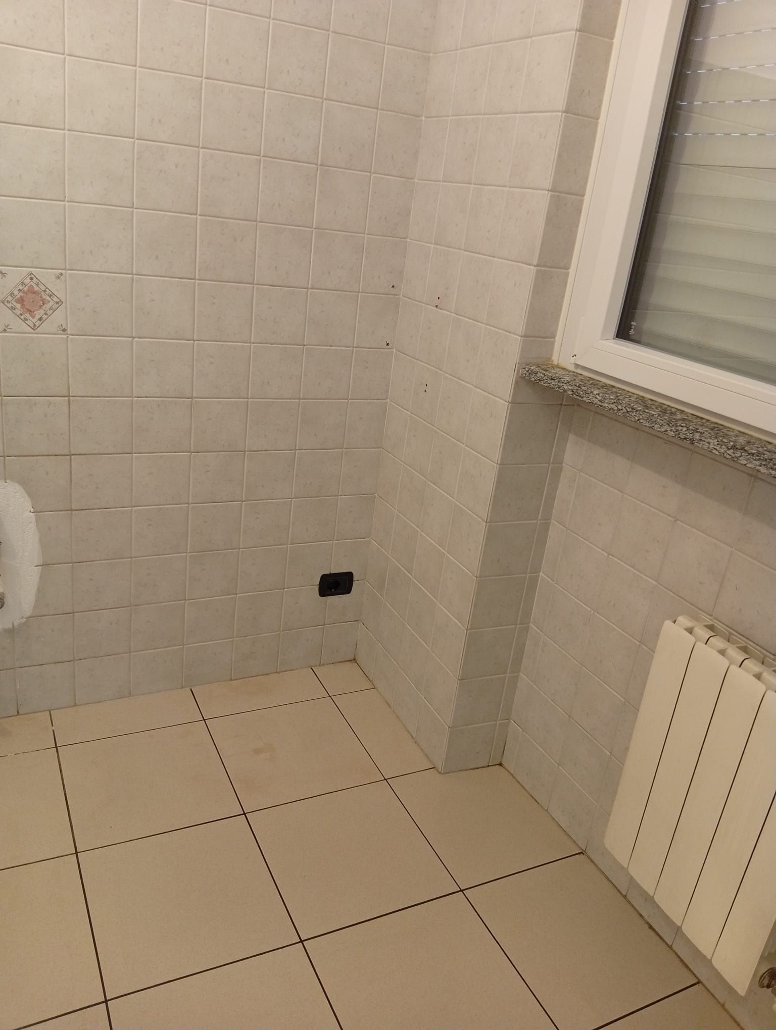 Angolo del bagno piastrellato e vuoto, con finestra e termosifone, che mostra una piccola presa a muro vicino al pavimento.