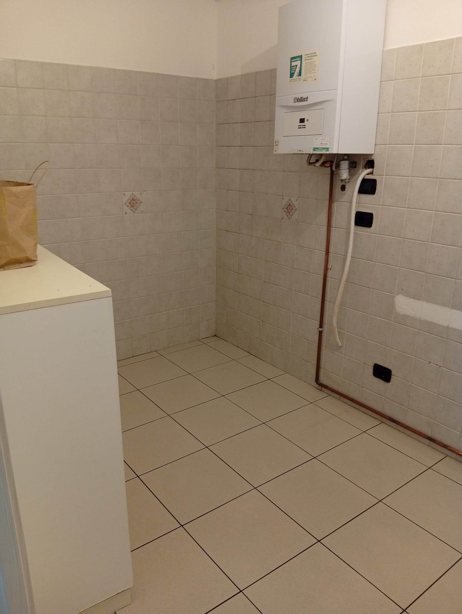 Locale di servizio vuoto piastrellato con caldaia a muro e armadietto bianco nell'angolo.