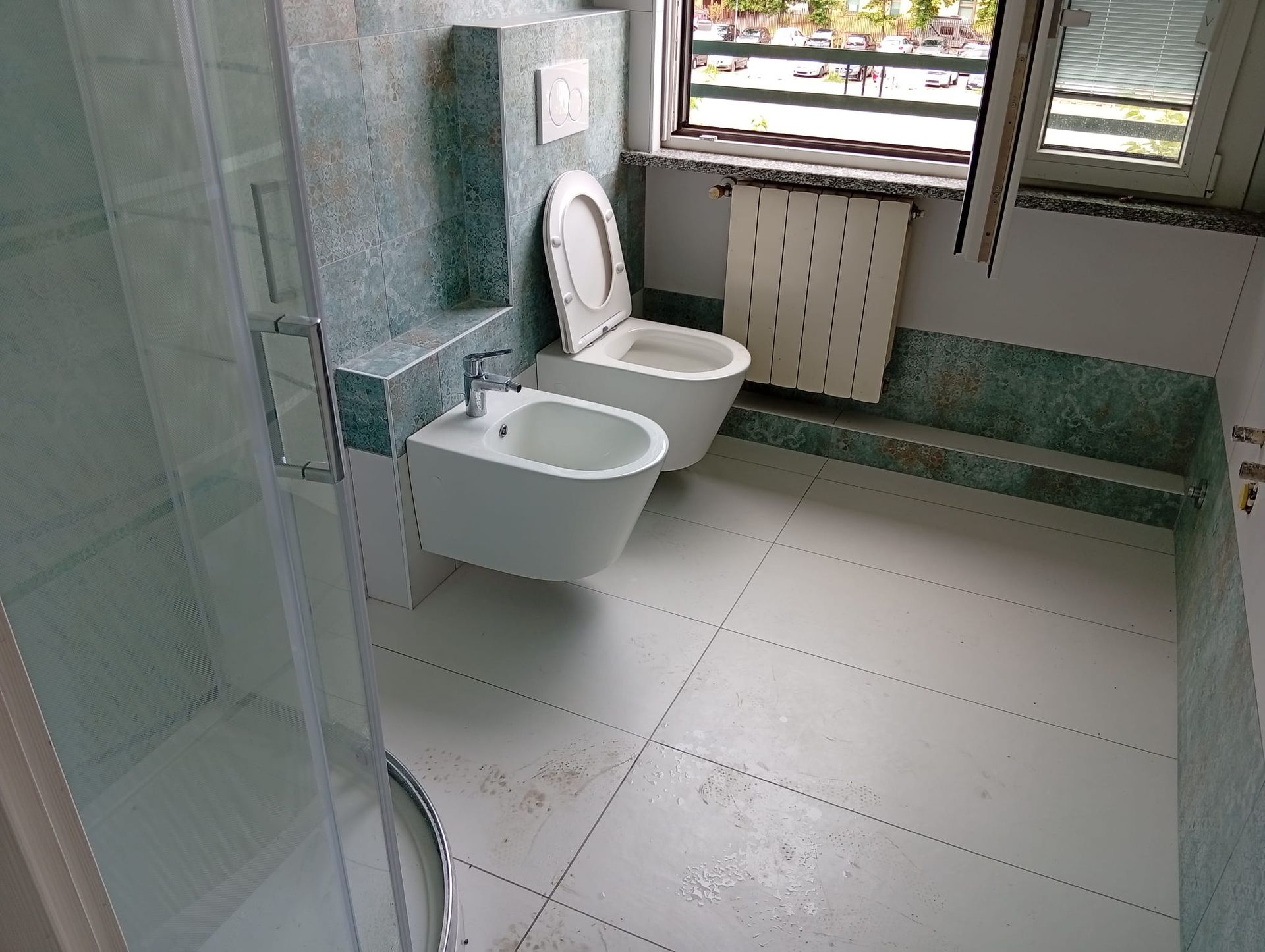 Bagno pubblico con WC a parete, bidet e pavimento piastrellato vicino a una finestra.