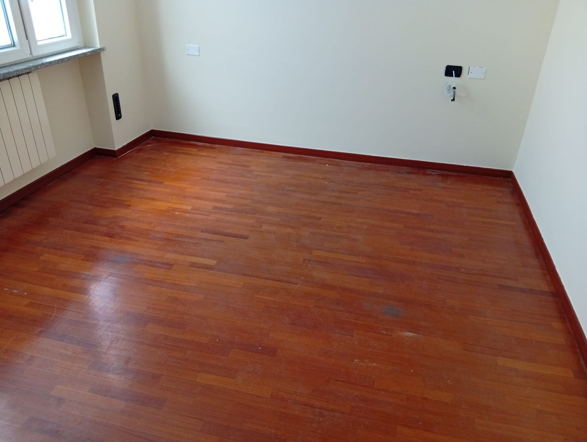 Stanza vuota con pavimento in legno rossastro, pareti bianche e un termosifone a finestra sulla sinistra.