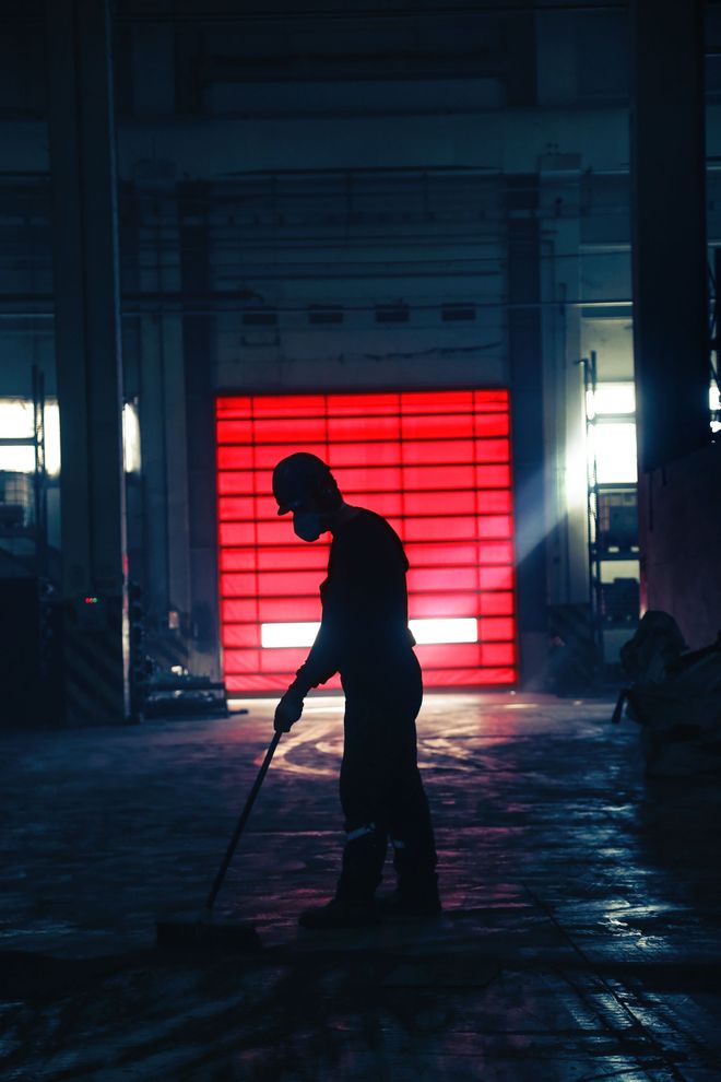 Silhouette di una persona che tiene un bastone in un magazzino poco illuminato, illuminata da un pannello retroilluminato rosso brillante.
