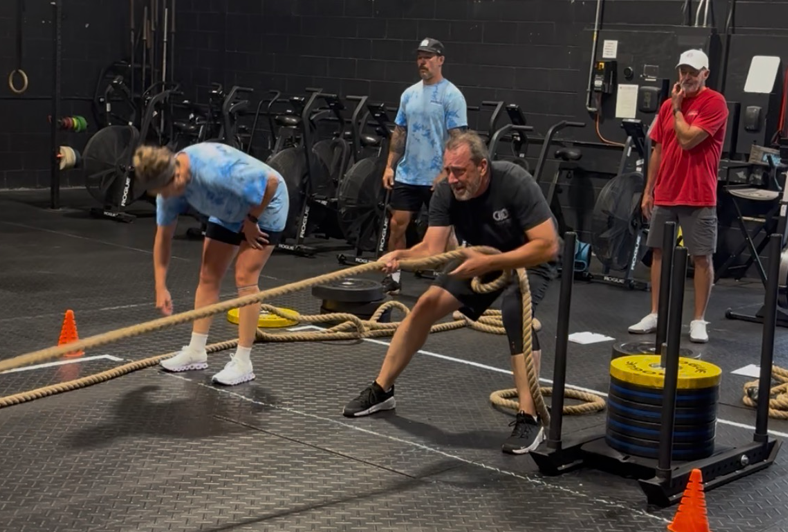 Two people exercising in a gym, one crouched with a resistance band facing the other.