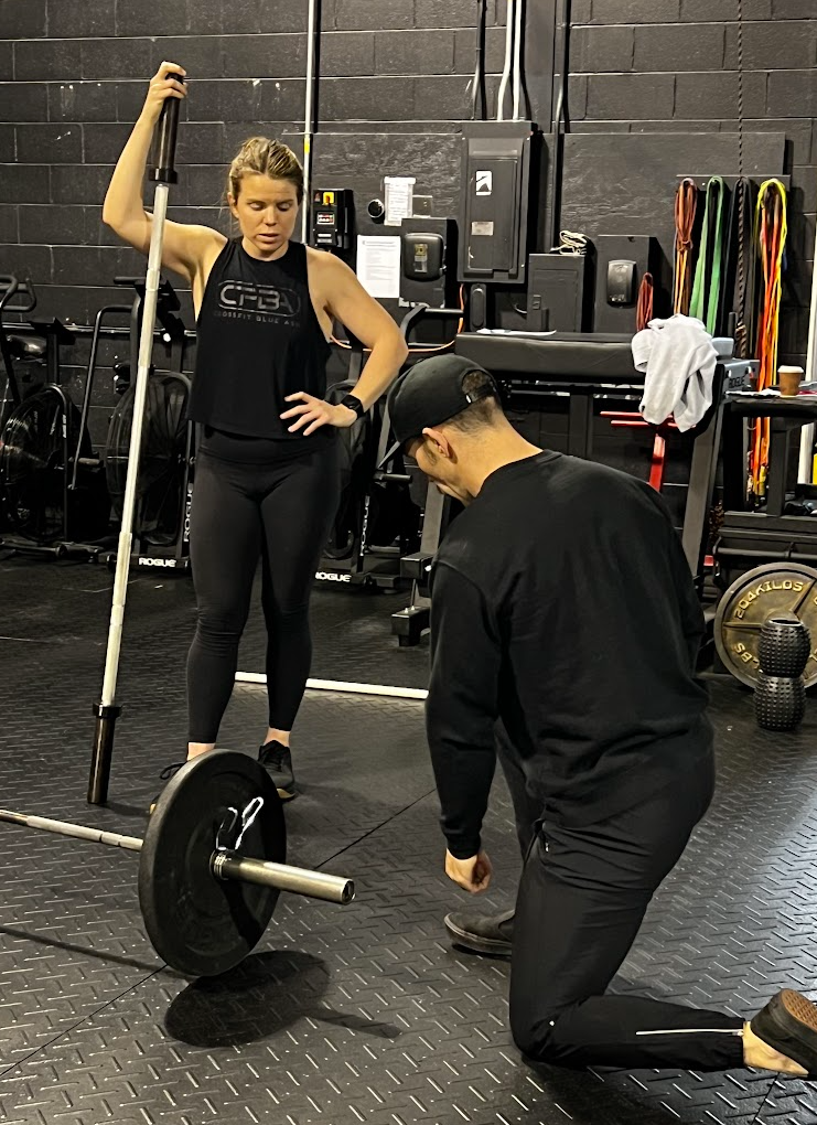 Two people in a gym adjusting a barbell on the floor, with one holding a staff nearby