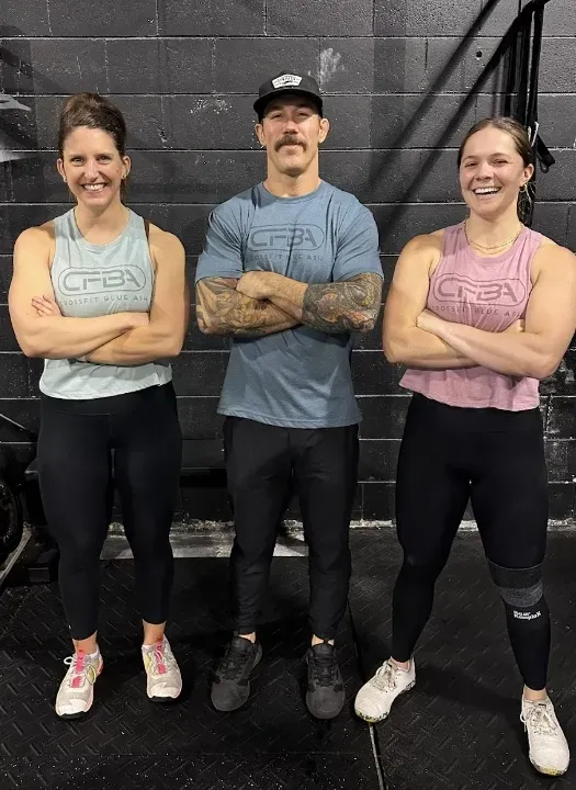 Three people standing with arms crossed in a gym, wearing workout clothes and smiling.