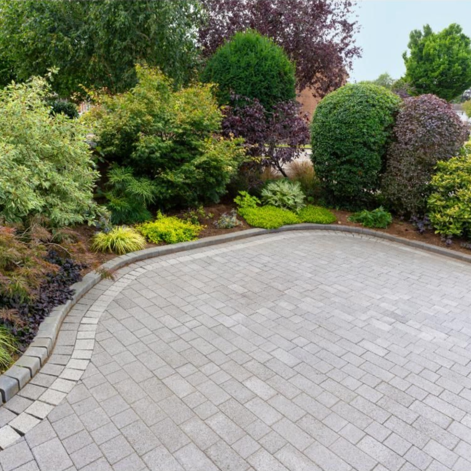 Curved gray paver driveway bordered by lush green shrubs and trees