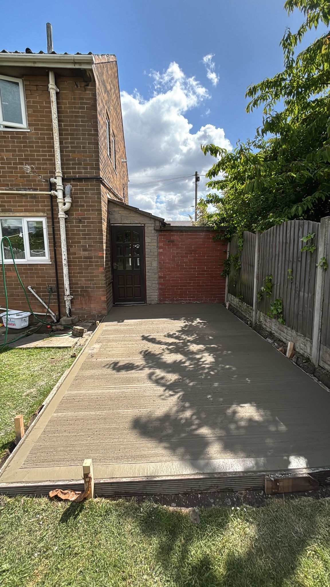 Concrete backyard patio beside a brick house, bordered by a fence and gate, with grass in the foreground.