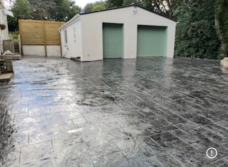 Small white garage with green doors beside a wet, gray stamped-concrete patio in a backyard