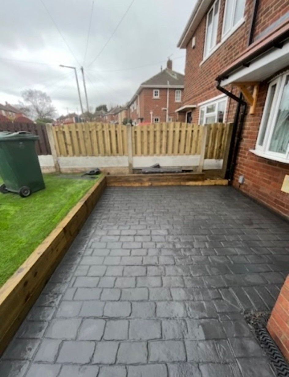 Paved side yard with brick house, wooden fence, green lawn, and a green bin on the left.