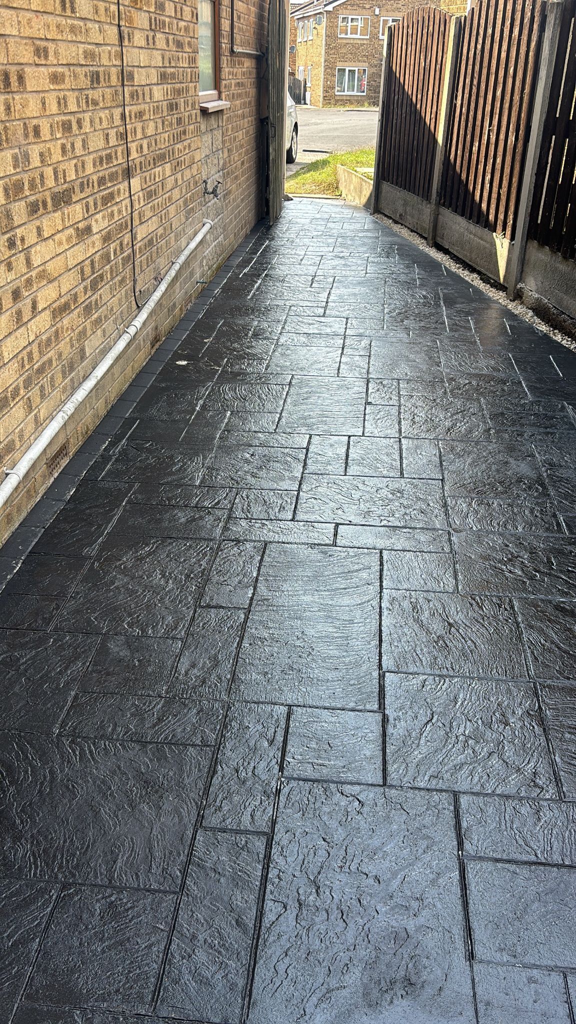 Narrow wet stone alleyway between a brick wall and wooden fence, leading to a bright opening.
