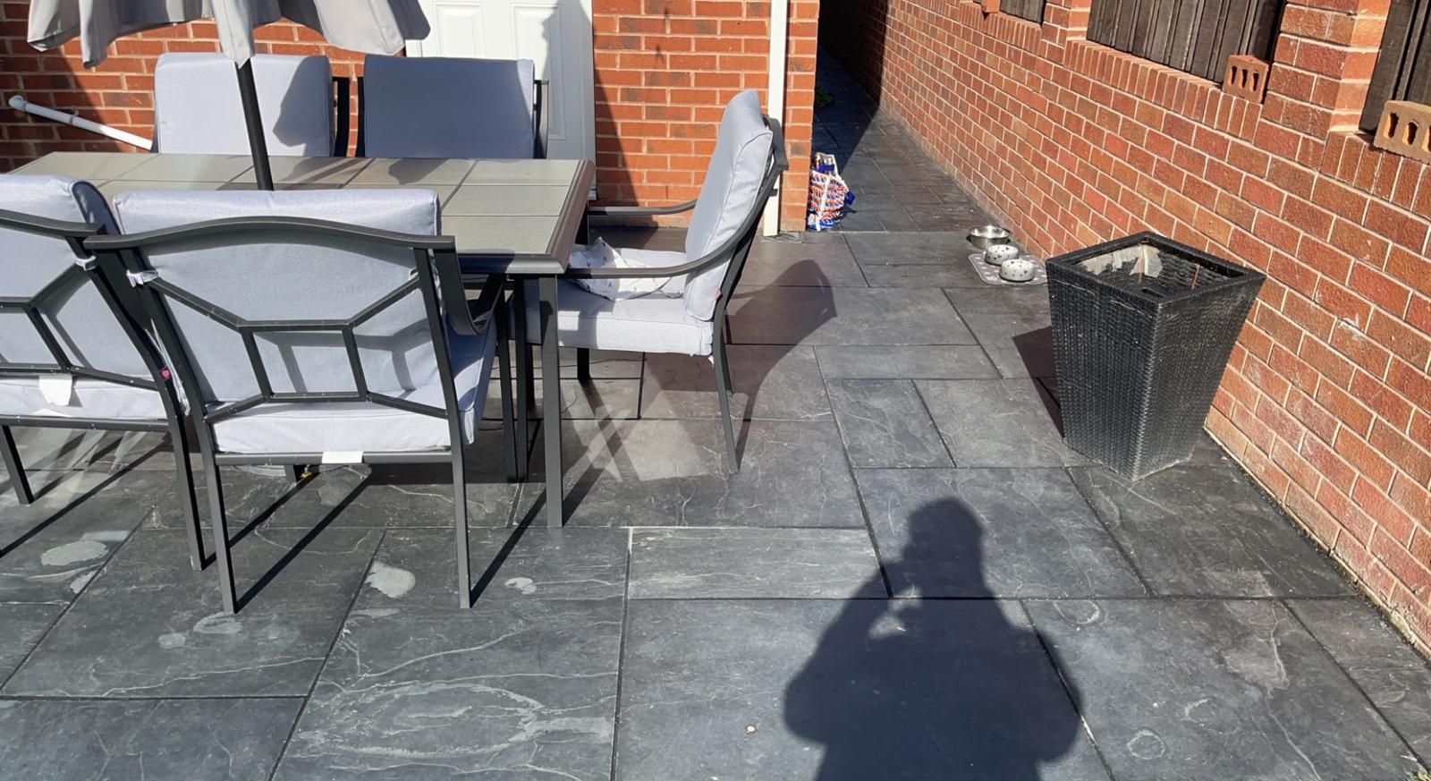 Outdoor patio with metal chairs and table on gray stone tiles beside a red brick wall, with a person’s shadow in foreground