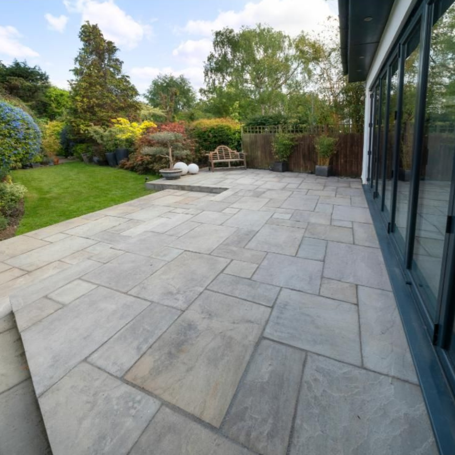 Large gray stone patio beside a house, bordered by a lush green garden and trees