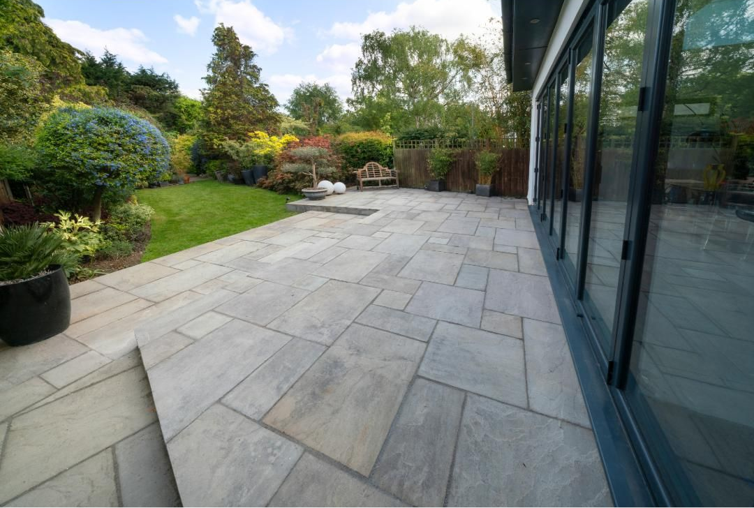 Stone patio beside glass doors, bordered by a lush green garden and trees.
