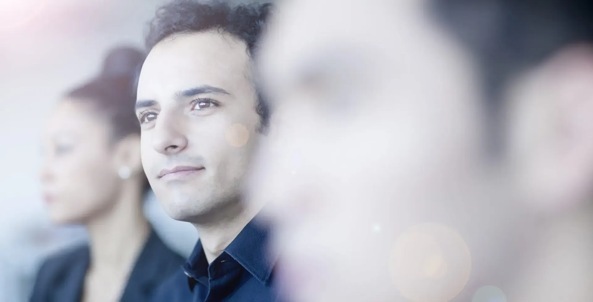 Man in focus, smiling, with two blurred people behind him. Bright light.