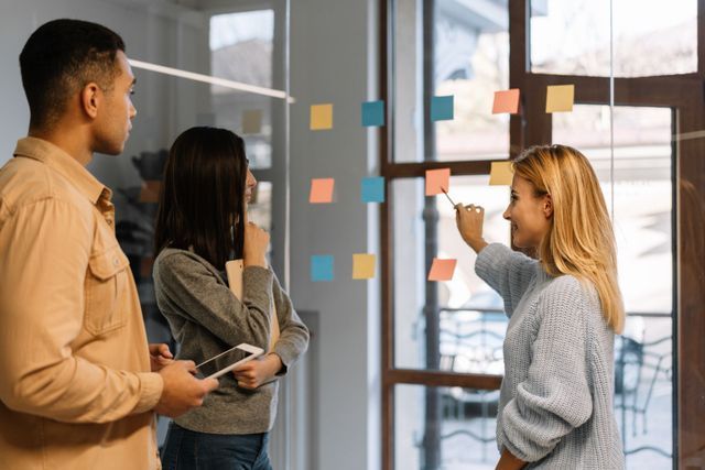 Three people in office, discussing sticky notes on a glass wall. One points with a pen, two look on.