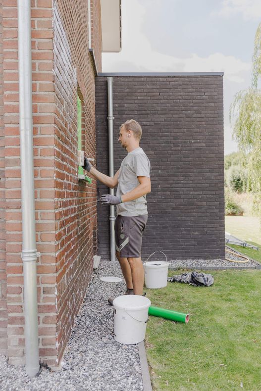 Een persoon schildert een bakstenen muur naast een huis met emmers en gereedschap op het gras.