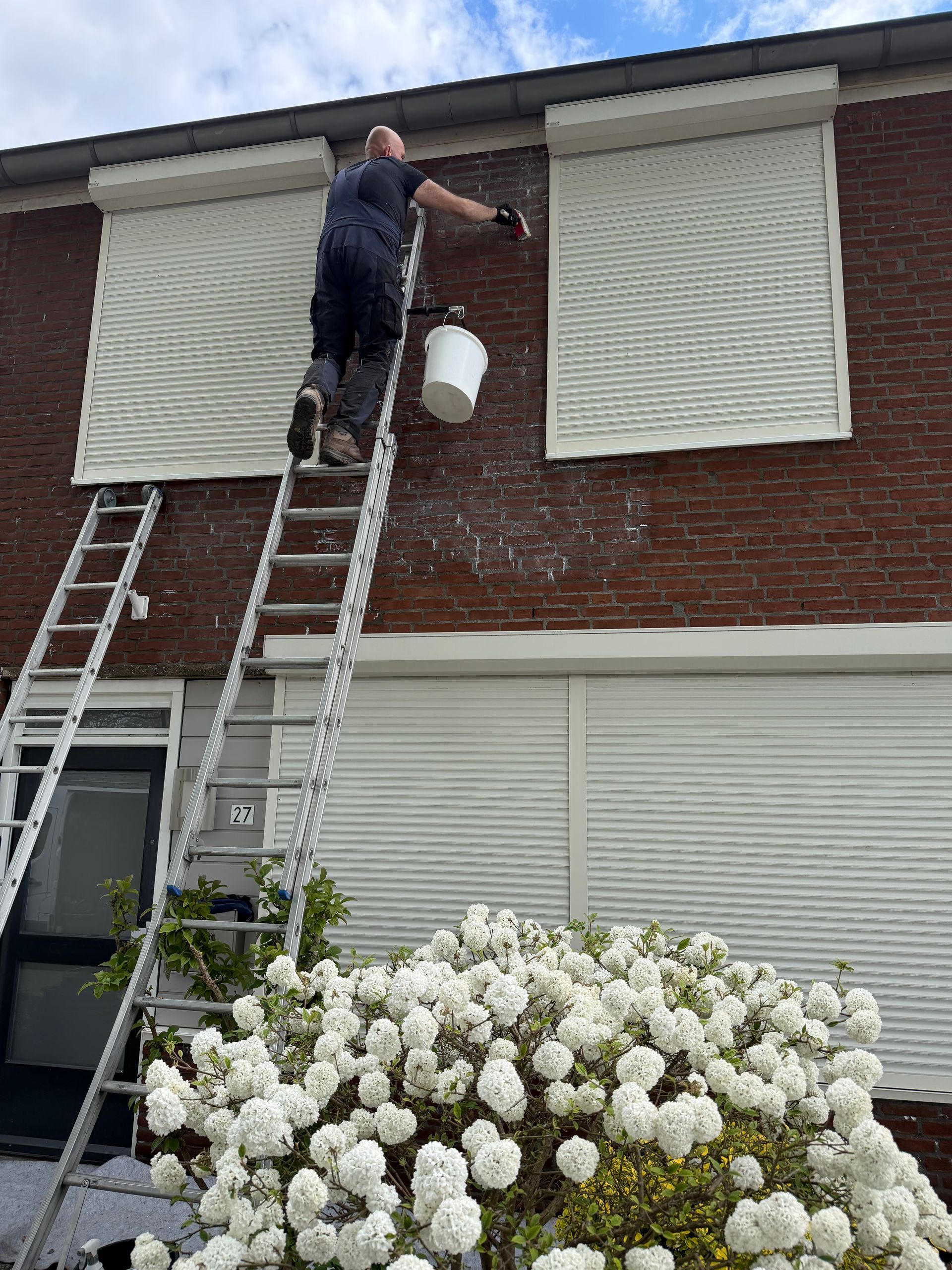 Een persoon reinigt met een hogedrukreiniger de bakstenen voorgevel van een huis; links op de foto is een ladder te zien.
