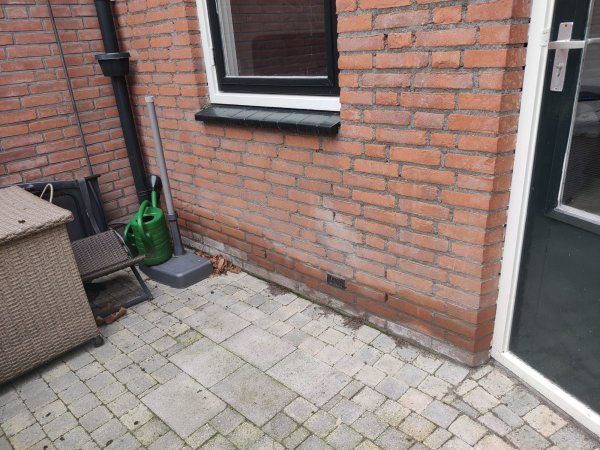 Detailopname van een gebarsten muurplint met rode verf, blootliggend bruin pleisterwerk en een afbrokkelende betonnen vloer.