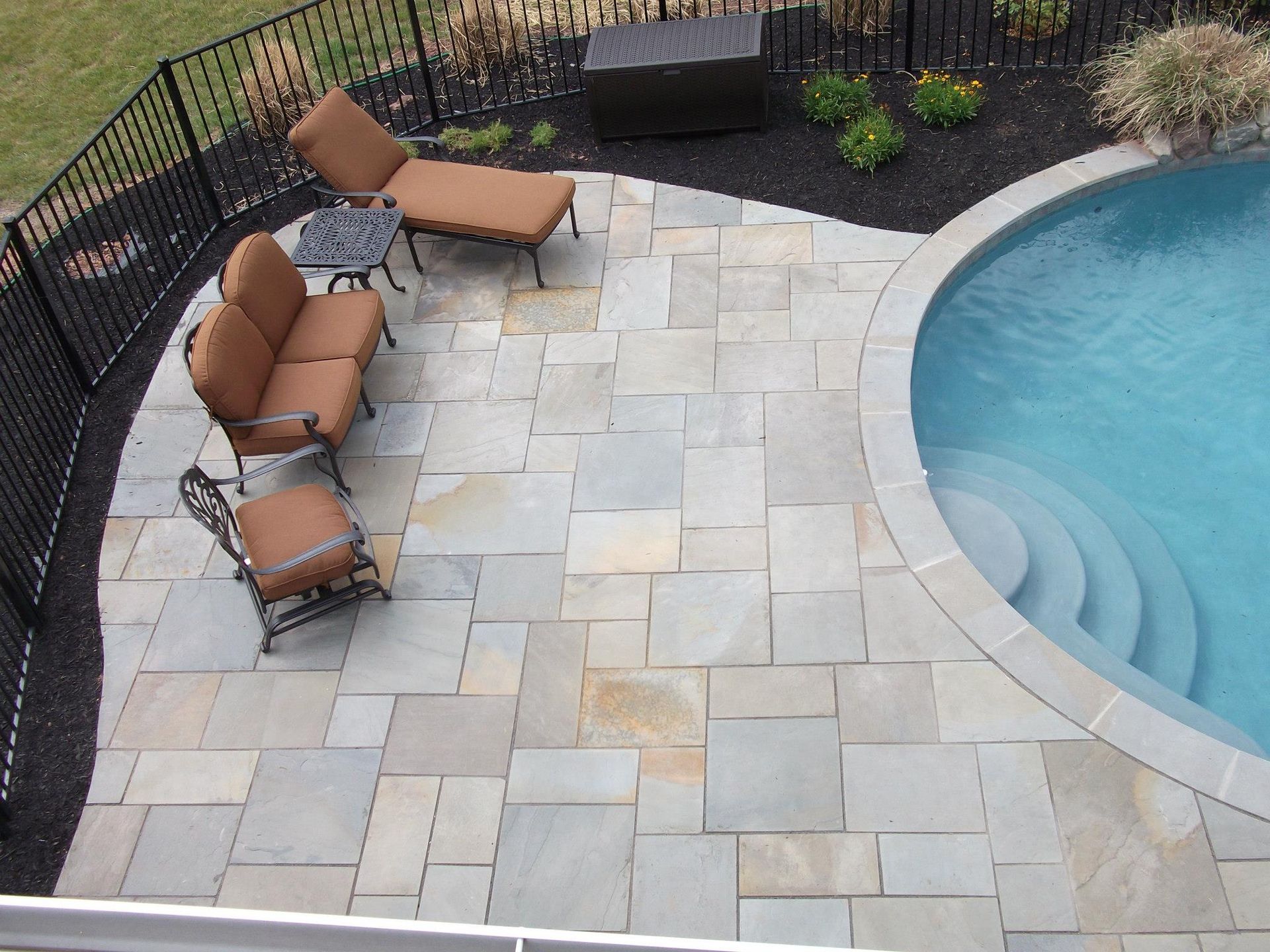 An aerial view of a stone patio with outdoor furniture next to a swimming pool with steps, framed by a black fence.