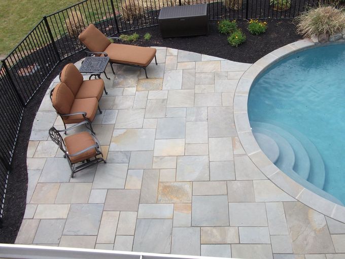An aerial view of a stone patio with outdoor furniture next to a swimming pool with steps, framed by a black fence.