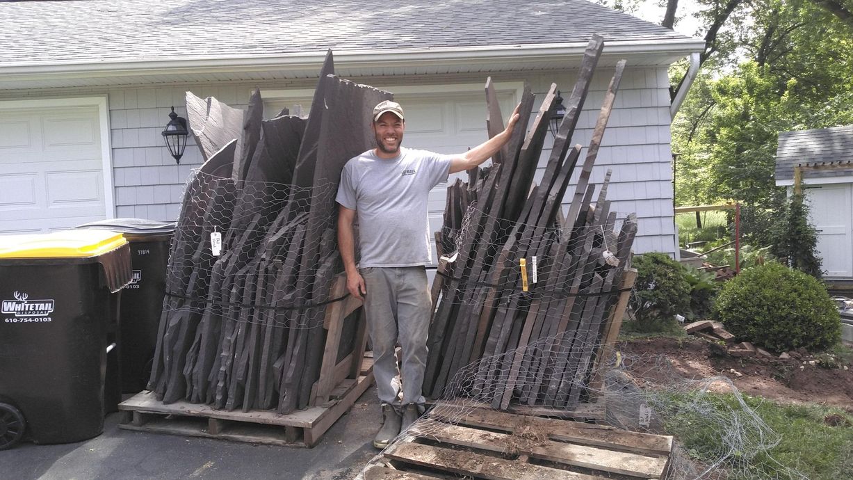 A person stands between two large, strapped pallets of dark, thin-cut stone veneer in a residential driveway.