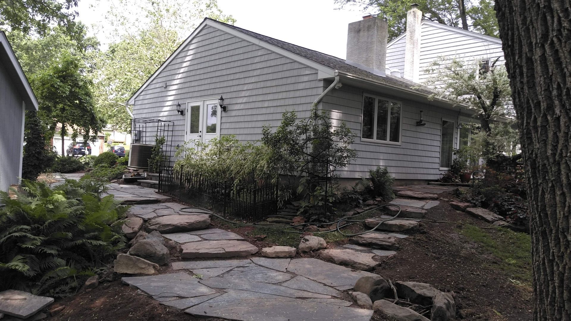 A gray-sided house with a chimney and white doors, surrounded by trees and a stone-paved path through a garden.