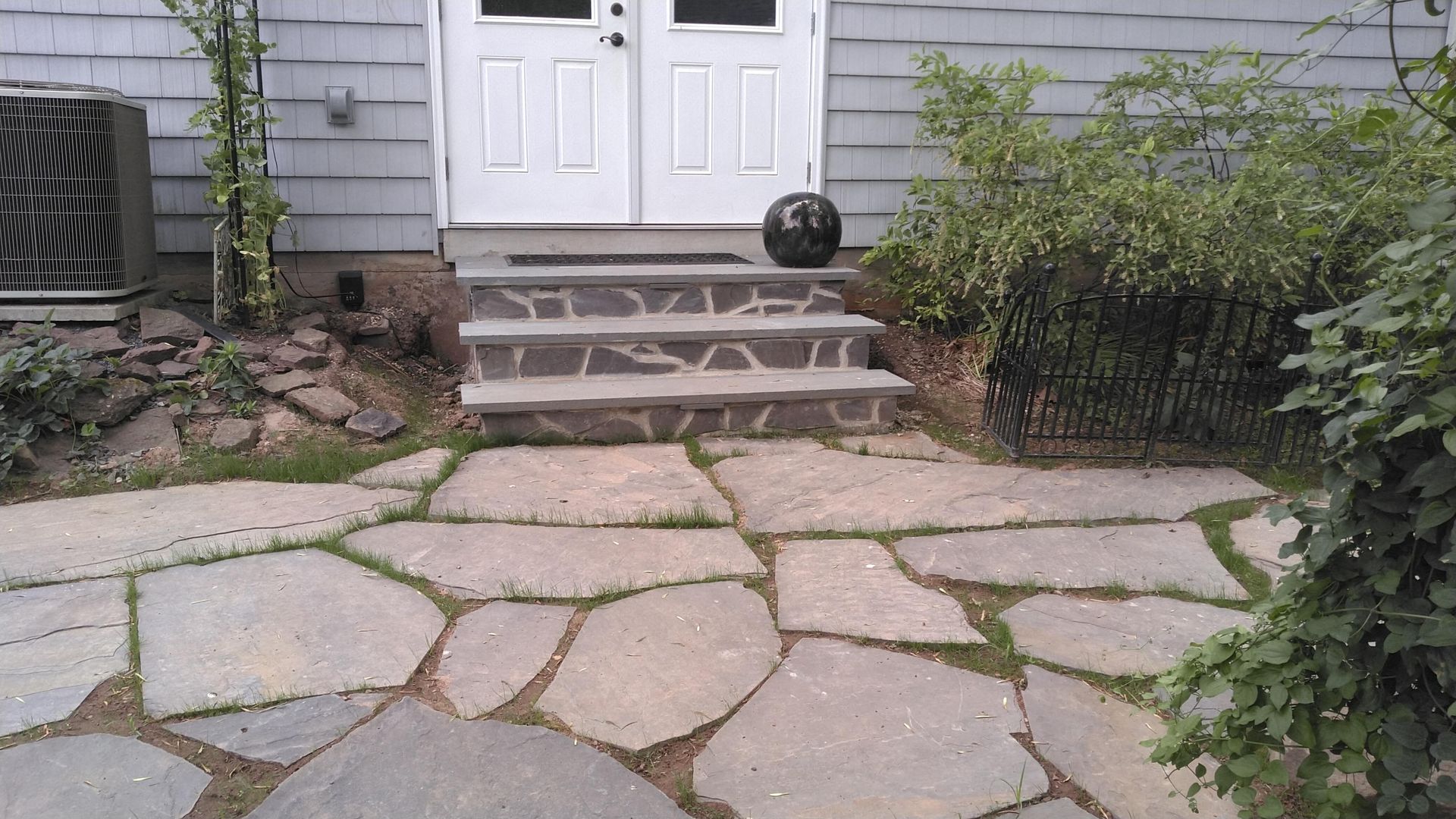 A stone patio leads to a short set of stone steps and double white doors on a gray-sided house.