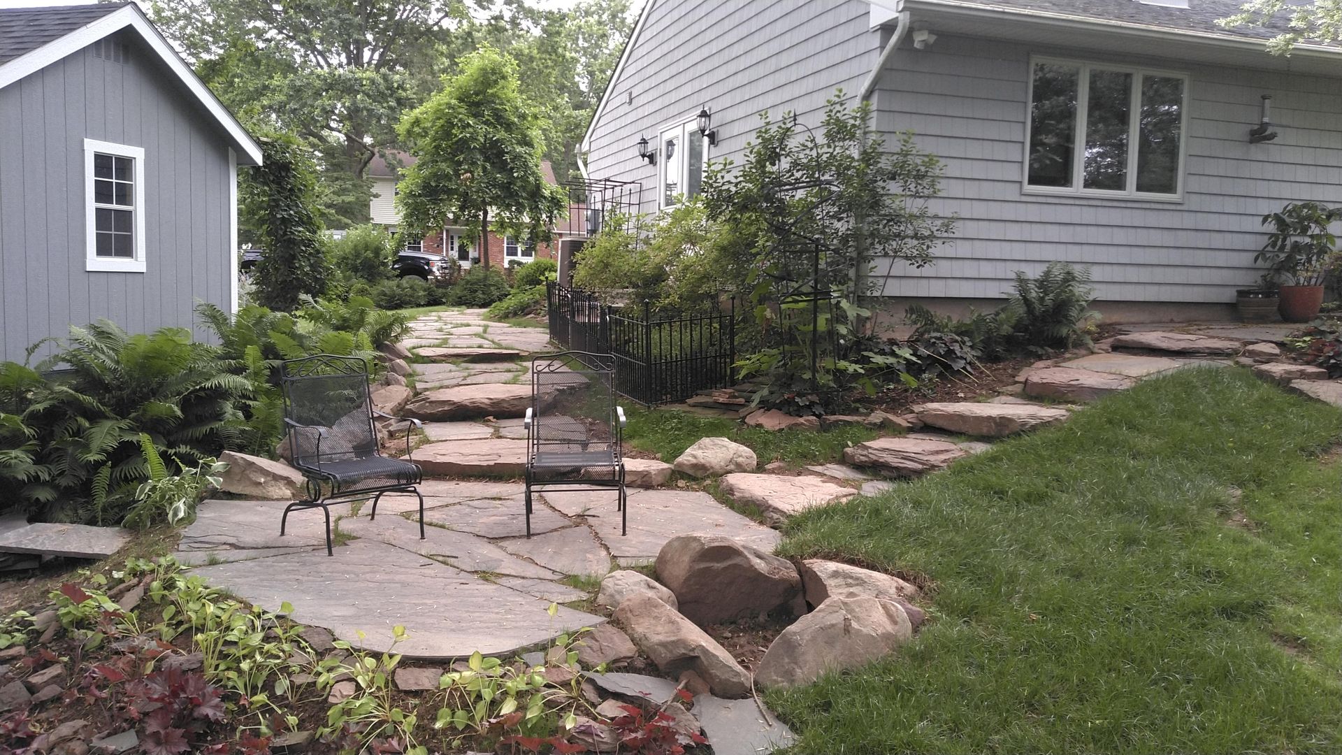 Two metal chairs sit on a stone patio in front of a house, surrounded by plants and garden paths.