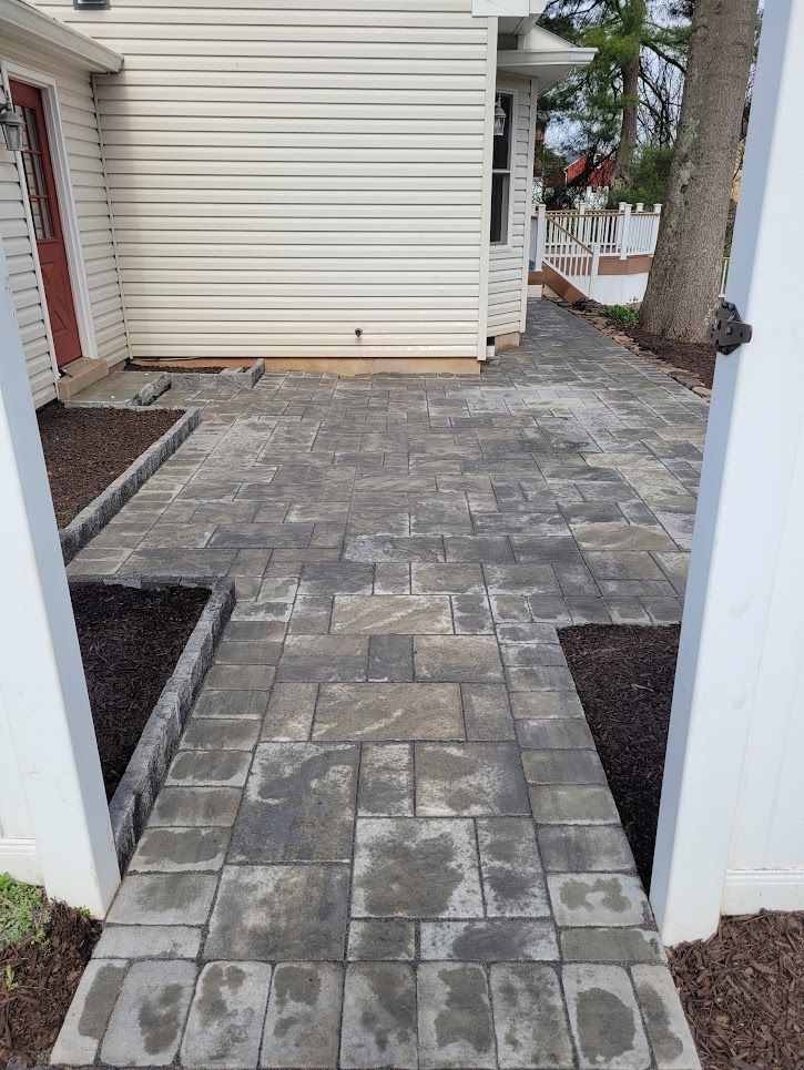 A gray stone paver walkway leads to the side entrance of a house with beige siding, flanked by mulch-filled garden beds.