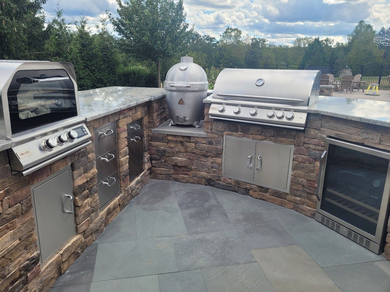 An outdoor kitchen featuring a grill, a ceramic smoker, and a small refrigerator built into a stone counter on a patio.