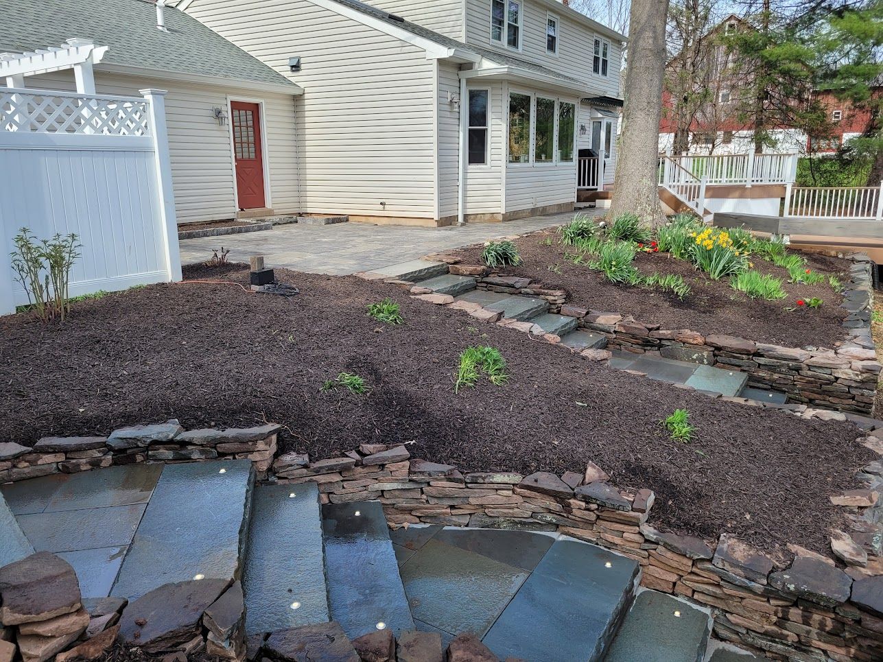 A backyard landscape with stone steps leading up to a mulched garden bed and a patio next to a two-story house.