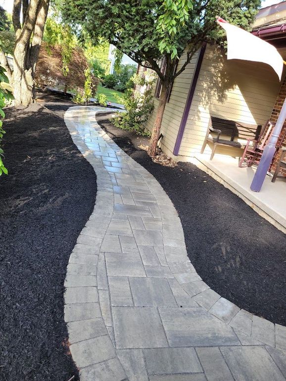 A gray stone walkway with a curved design, bordered by fresh dark mulch, leads alongside a house with a shaded porch.