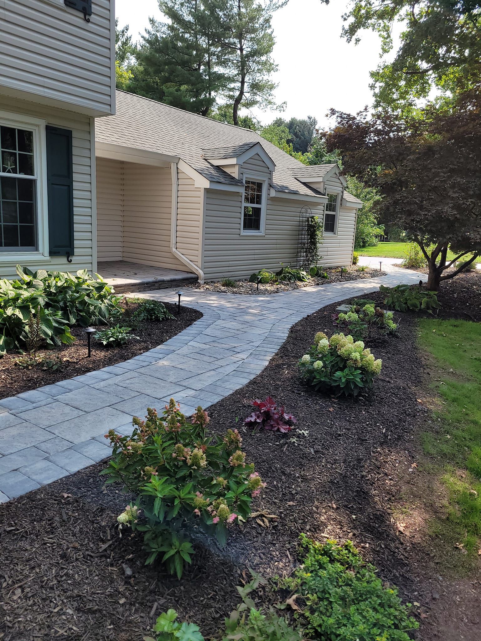 A light-colored house with a stone walkway leading to the entrance, surrounded by mulched garden beds and greenery.