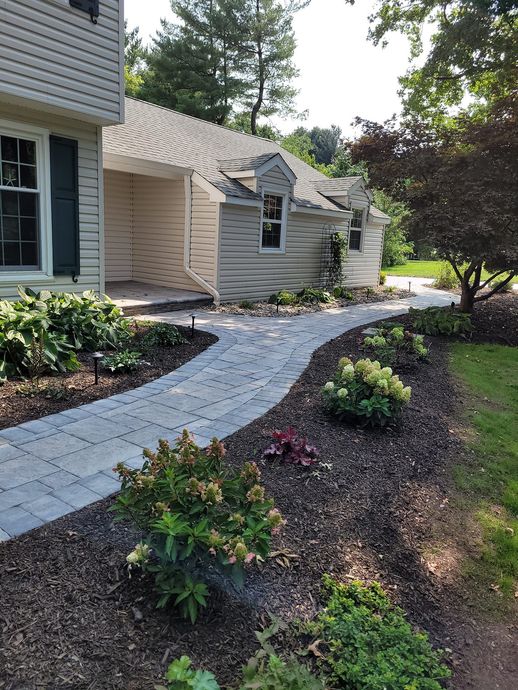 A paved stone walkway leads through a landscaped yard to the entrance of a light-colored house with dark shutters.