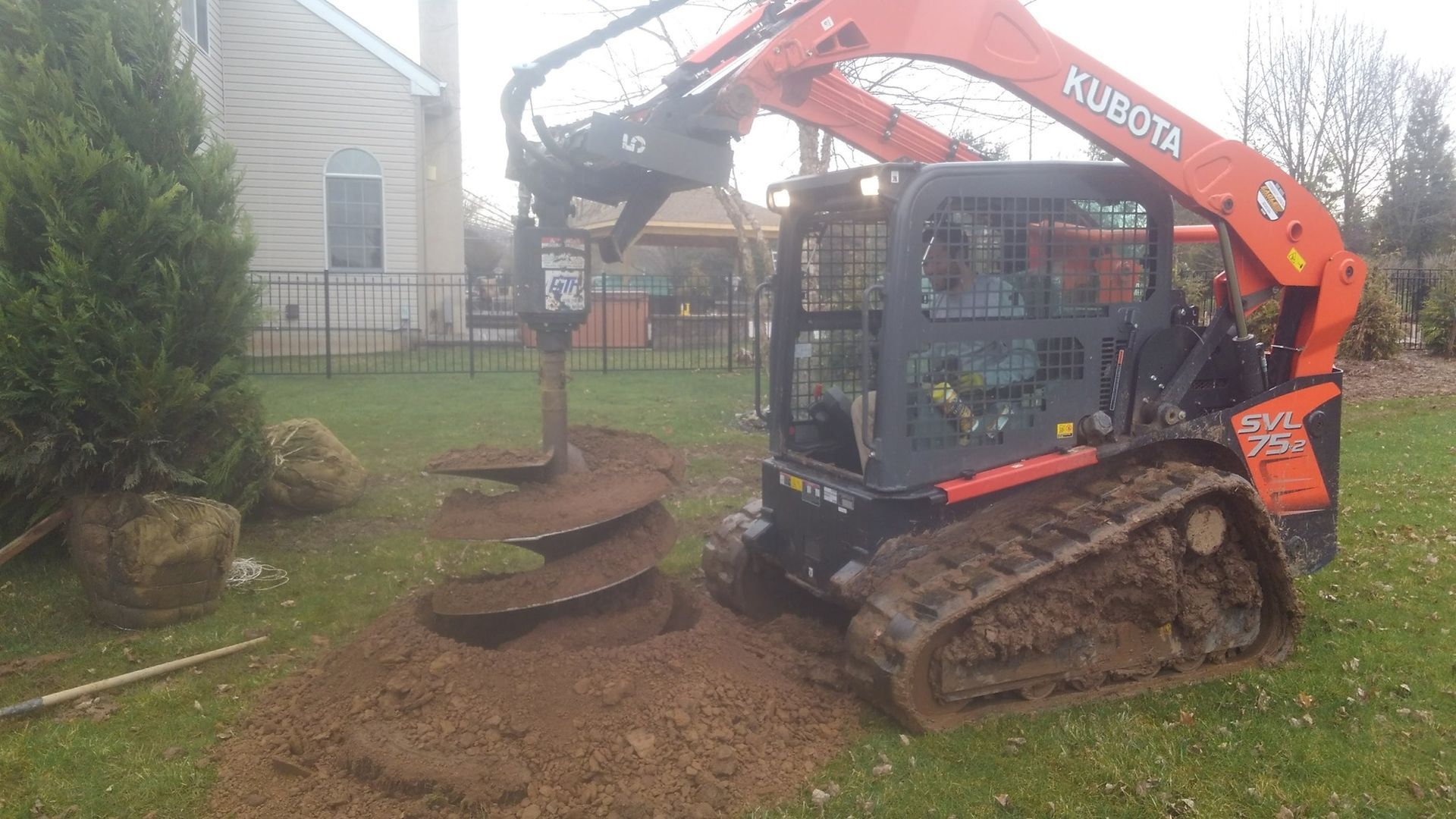 An orange Kubota skid-steer loader uses an attached earth auger to drill a hole in a residential backyard.