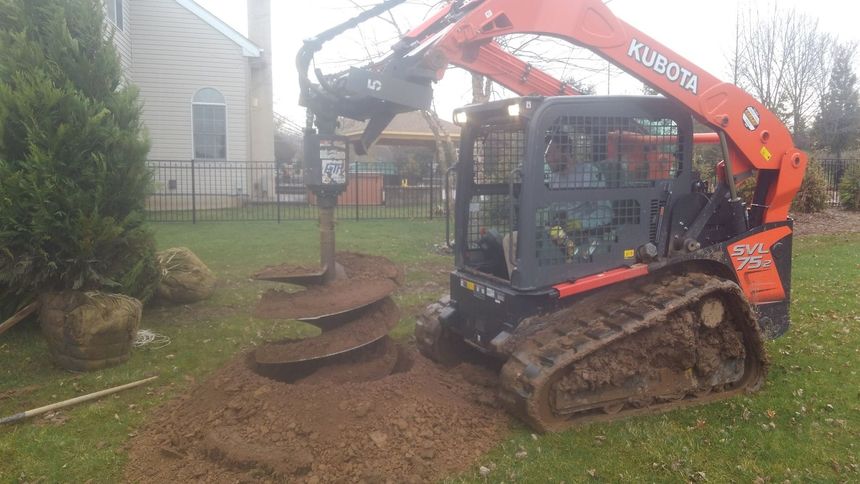 An orange Kubota skid-steer loader uses an attached auger to drill a hole in a residential grassy yard.