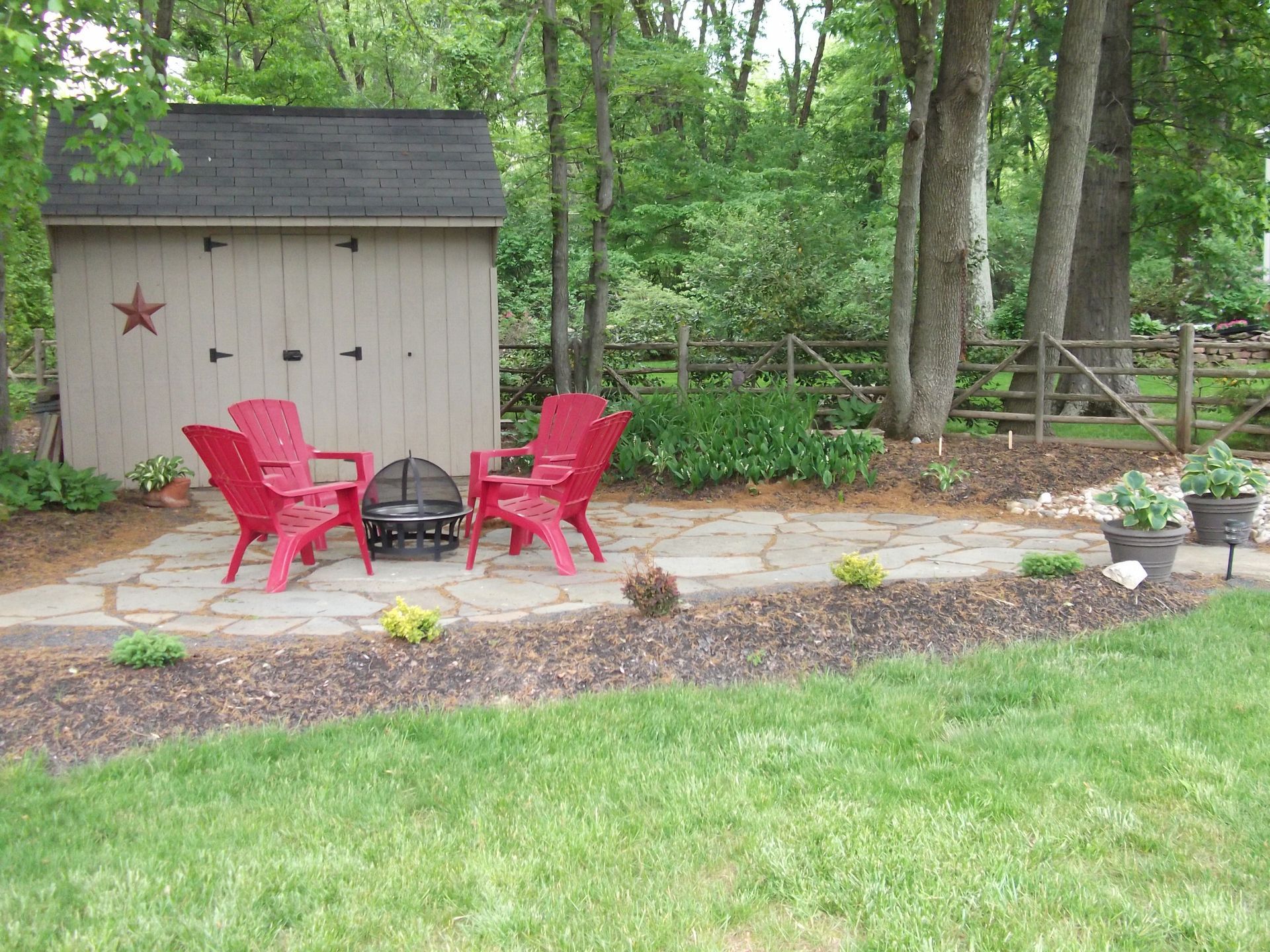 Backyard patio with red chairs, fire pit, stone path, and a small shed surrounded by trees