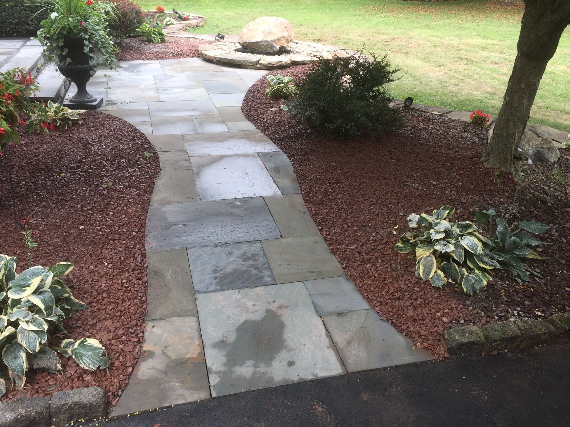 A stone walkway winds through a garden bed with red mulch, hosta plants, shrubs, and a large decorative boulder.
