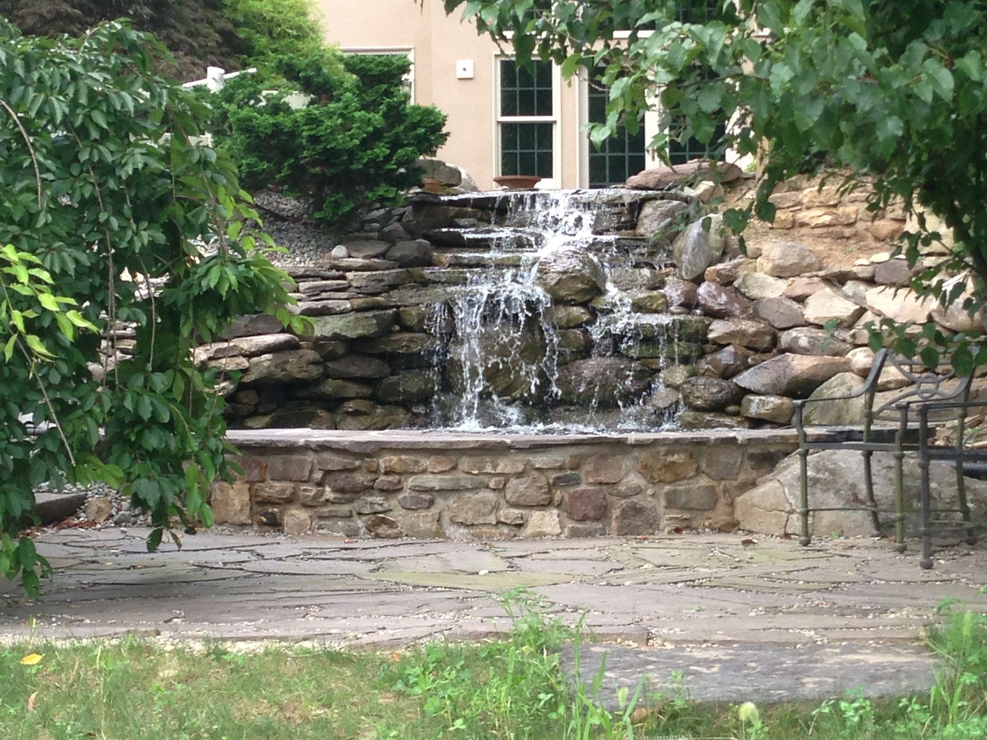 Stone waterfall fountain in a garden with rocks, greenery, and a stone patio