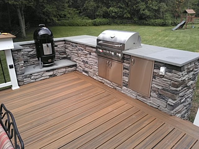 A stone outdoor kitchen with a built-in stainless steel grill, cabinets, and a charcoal smoker on a wooden deck.