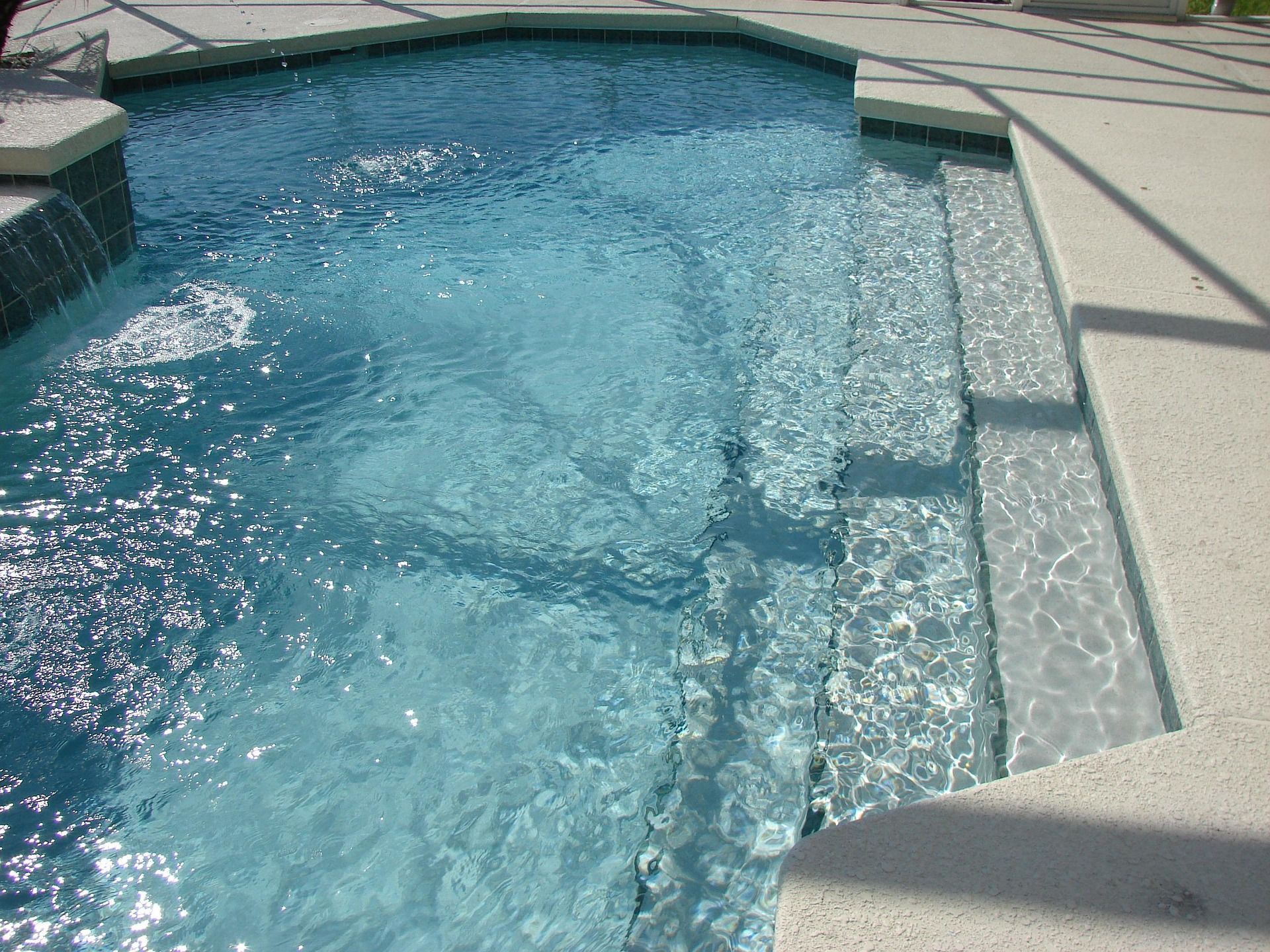 Una piscina di un azzurro cristallino con scalini e pavimentazione in cemento bianco, riparata da un ombrellone.