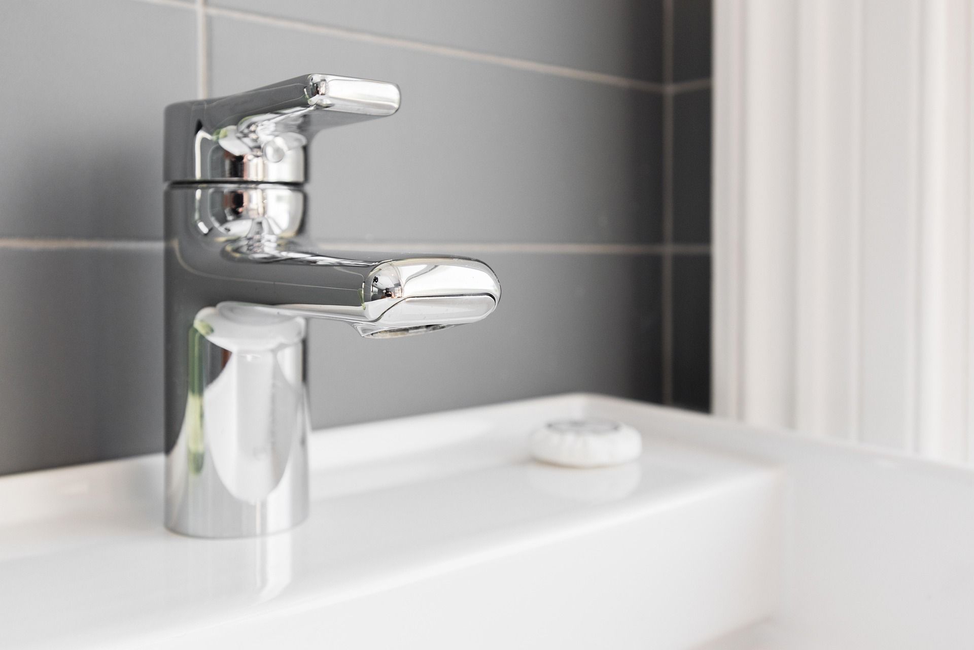 Un rubinetto cromato su un lavabo bianco del bagno, sullo sfondo di pareti rivestite di piastrelle grigie, con una piccola saponetta nelle vicinanze.