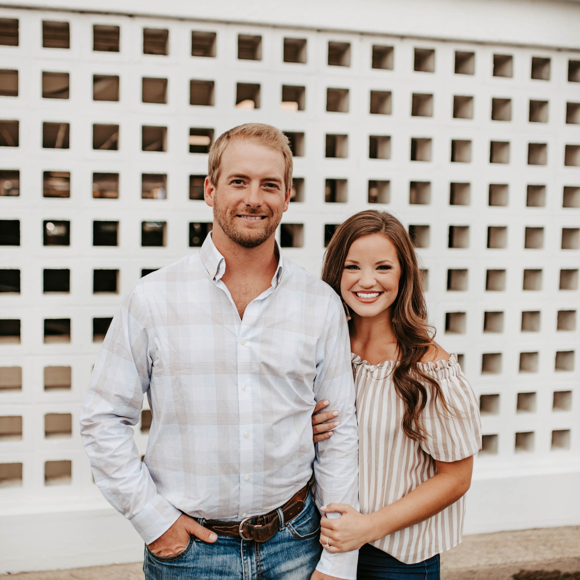 Man and woman pose in front of a white patterned wall, smiling. Man in blue-striped shirt; woman in striped top.