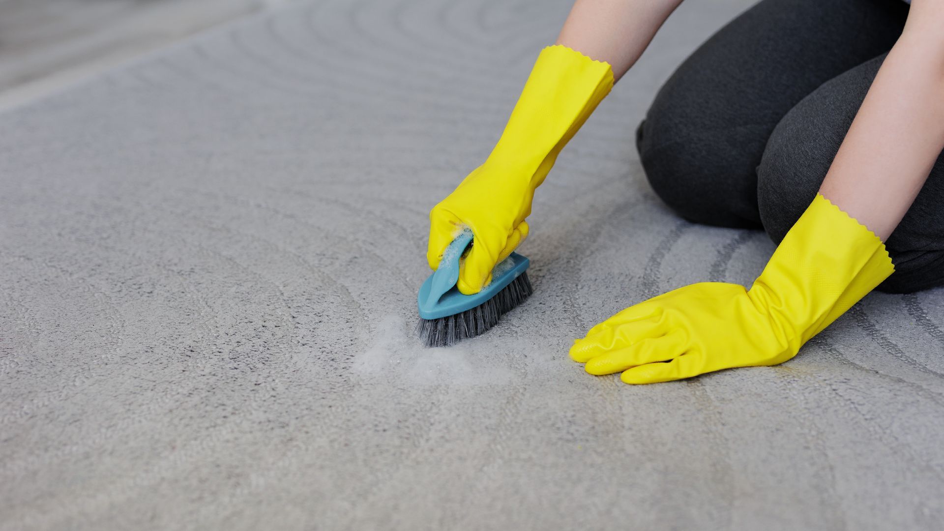 A person wearing yellow gloves is cleaning a carpet with a brush.