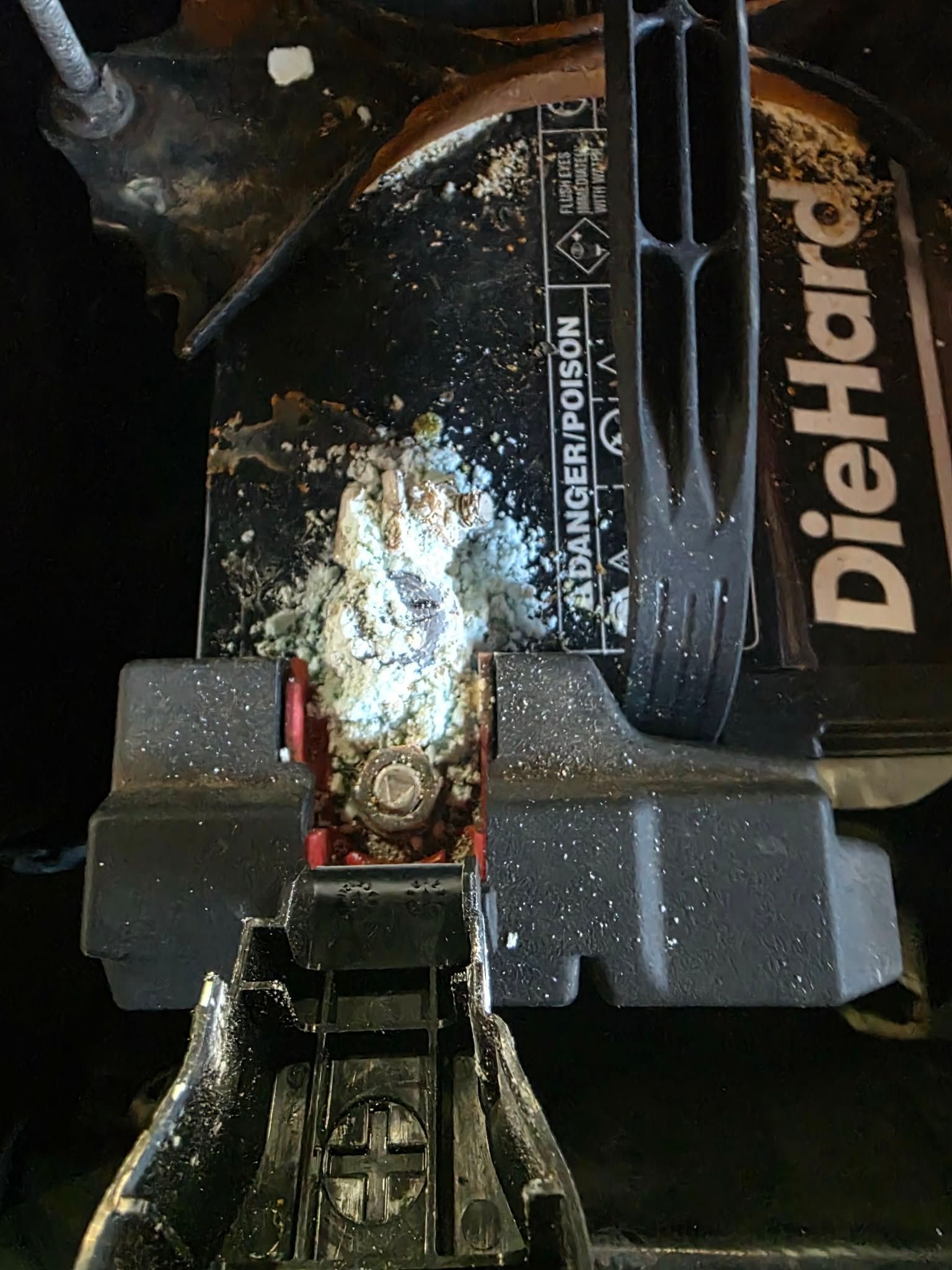 A car battery covered in white corrosion, with a black hold-down bracket and warnings visible. | Seth's Auto Care