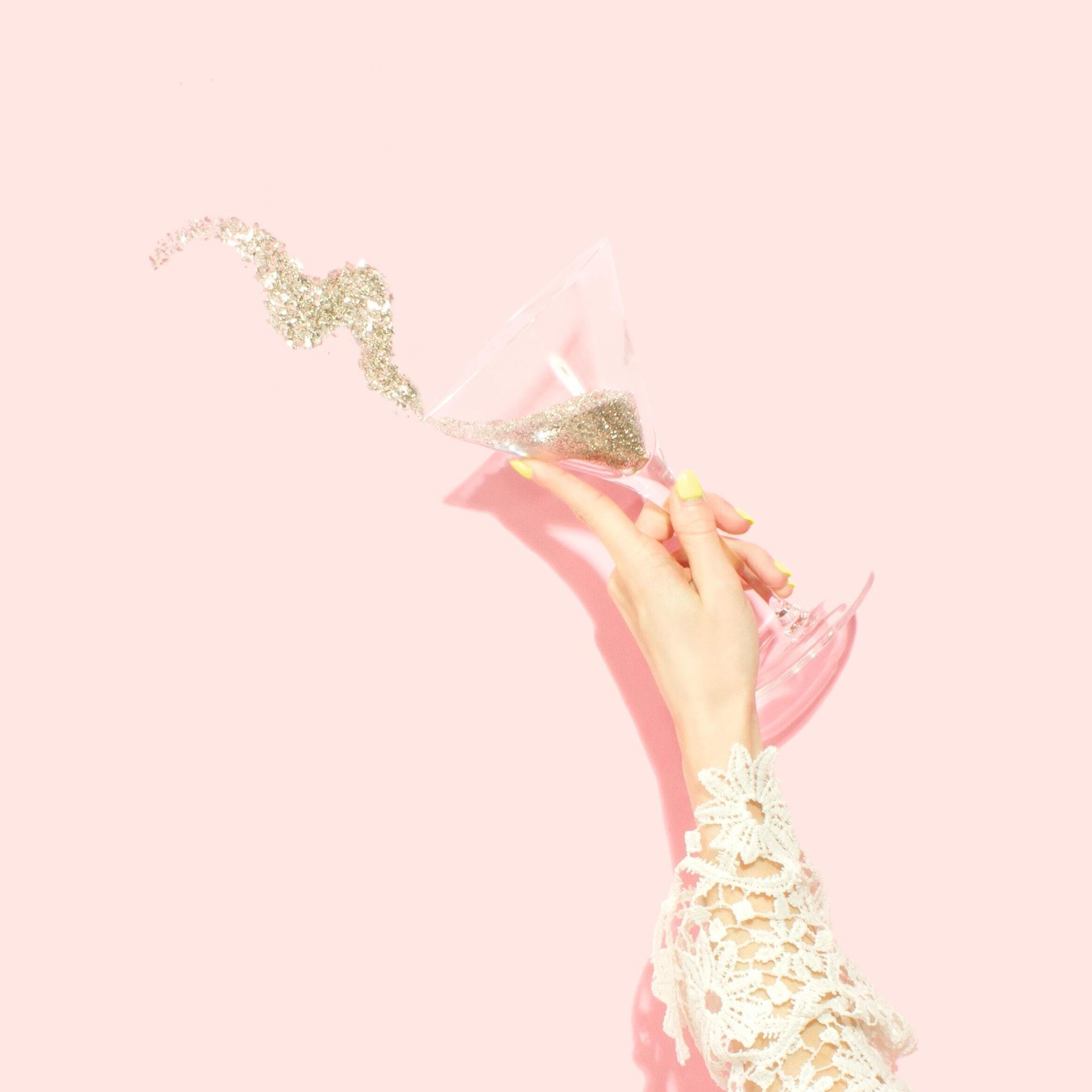 Hand holding a martini glass, pouring glitter on a pink background.