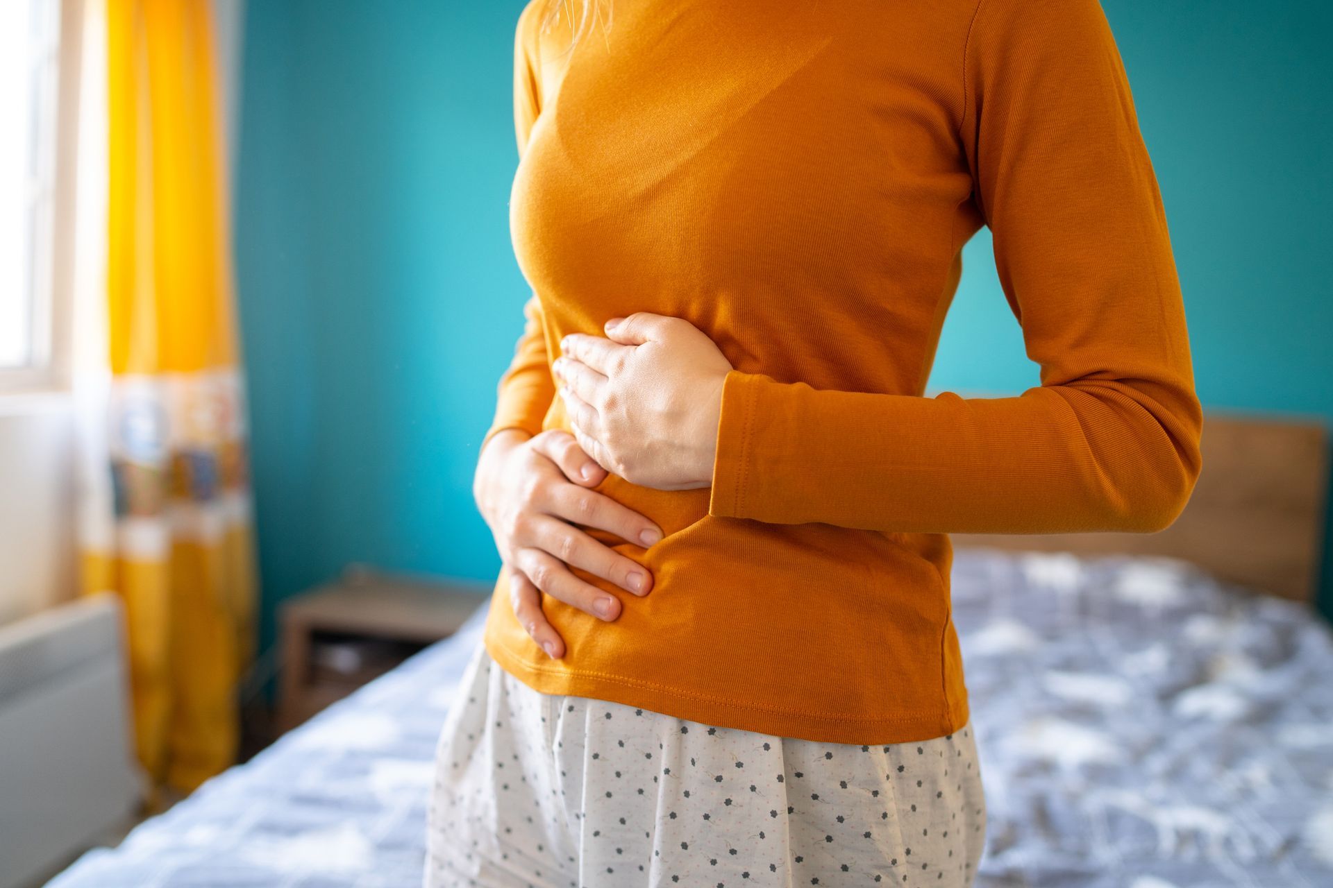 A woman is holding her stomach in pain while standing in front of a bed.